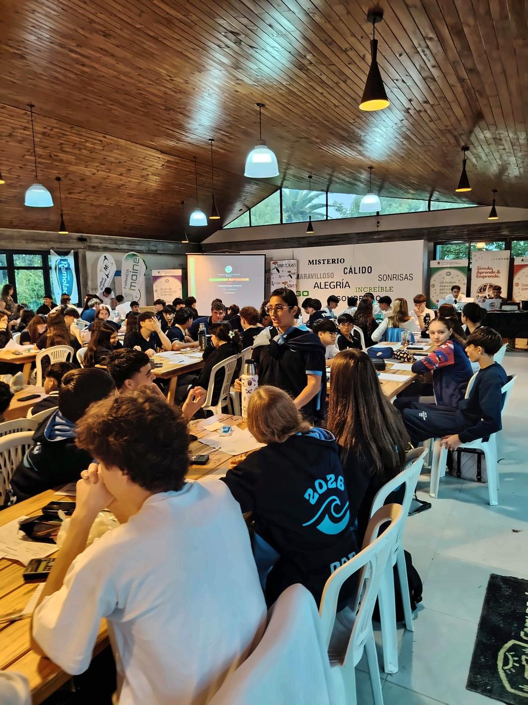 Salón lleno de equipos durante una instancia del certamen, con una docente acompañando a los estudiantes